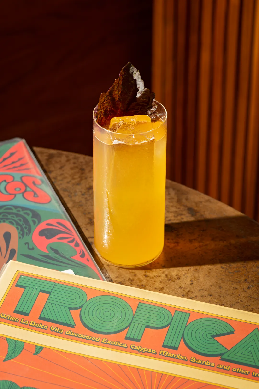 A tall golden cocktail with shiso beside a Tropicalia record